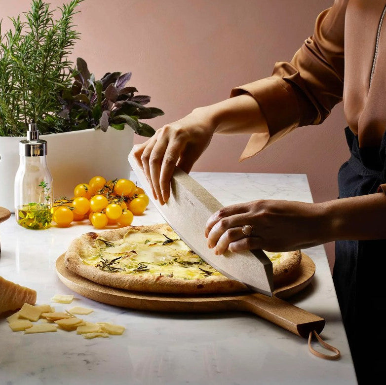 Person preparing a pizza on a wooden board with ingredients and plants in the background